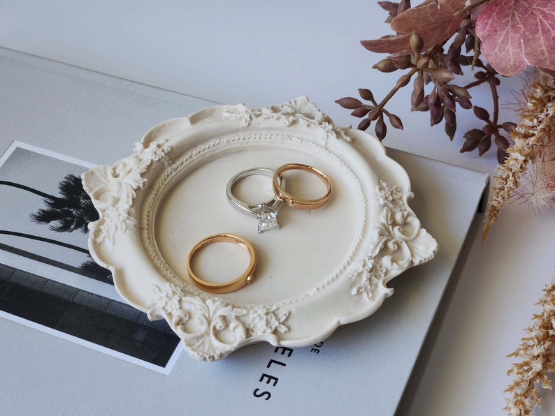 Puede incluir: Plato de cer&aacute;mica blanca para anillos con detalles ornamentados, que presenta tres anillos: una banda dorada, una banda plateada y una banda plateada con un diamante de talla cuadrada.