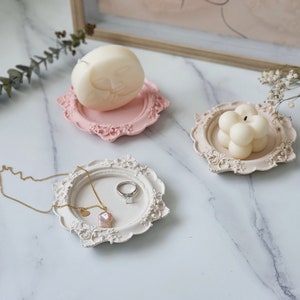 Puede incluir: Tres bandejas decorativas con bordes adornados, cada una con una vela diferente. La primera bandeja es rosa y contiene una vela blanca con forma de cara. La segunda bandeja es blanca y contiene un anillo y un colgante rosa. La tercera bandeja es blanca y contiene una vela blanca de forma redonda.
