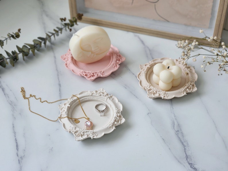 Puede incluir: Tres bandejas decorativas con bordes adornados, cada una con una vela diferente. La primera bandeja es rosa y contiene una vela blanca con forma de cara. La segunda bandeja es blanca y contiene un anillo y un colgante rosa. La tercera bandeja es blanca y contiene una vela blanca de forma redonda.