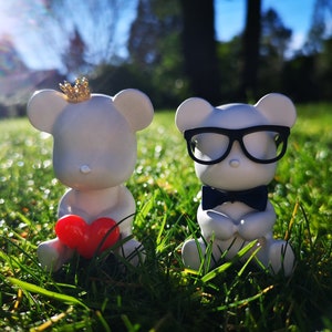 May include: Two white ceramic teddy bears, one wearing a gold crown and holding a red heart, the other wearing black glasses and a black bow tie, sitting in green grass.