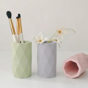 May include: Three geometric, pastel-coloured pencil holders. One is light green and holds makeup brushes. Another is grey with white flowers. The third is pink and lying on its side. These decorative holders are ideal for a desk or vanity.