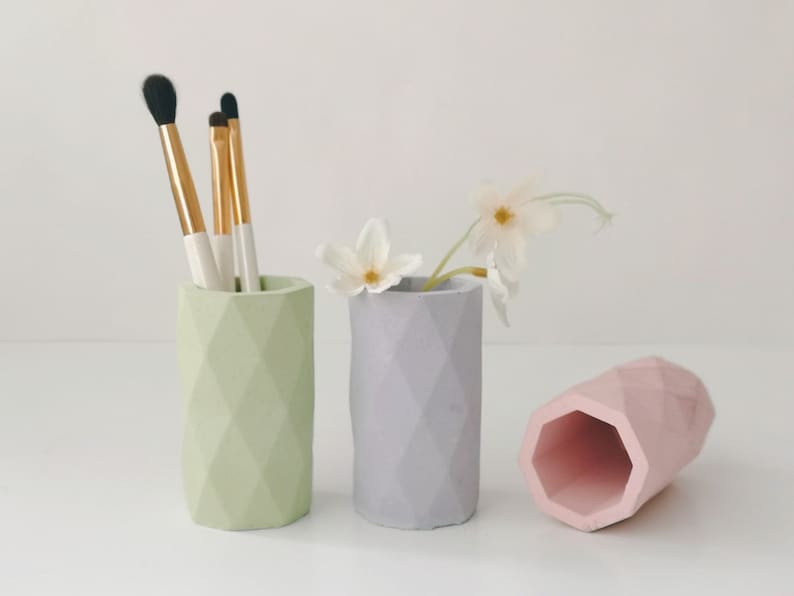 May include: Three geometric, pastel-colored pencil holders. One is light green and holds makeup brushes. Another is gray with white flowers. The third is pink and lying on its side. These decorative holders are ideal for a desk or vanity.