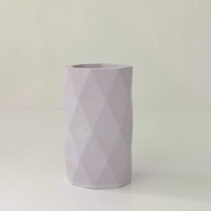 May include: A light purple, geometric vase with a faceted design. The cylindrical vase has a wide opening and is made of a solid material. The vase is a soft, muted color and stands on a white surface.