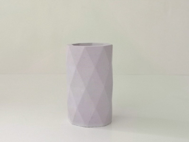 May include: A light purple, geometric vase with a faceted design. The cylindrical vase has a wide opening and is made of a solid material. The vase is a soft, muted color and stands on a white surface.