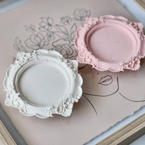 Puede incluir: Dos bandejas redondas decorativas, ornamentadas, blancas y rosas, con un marco y un dise&ntilde;o floral elevado y detallado.