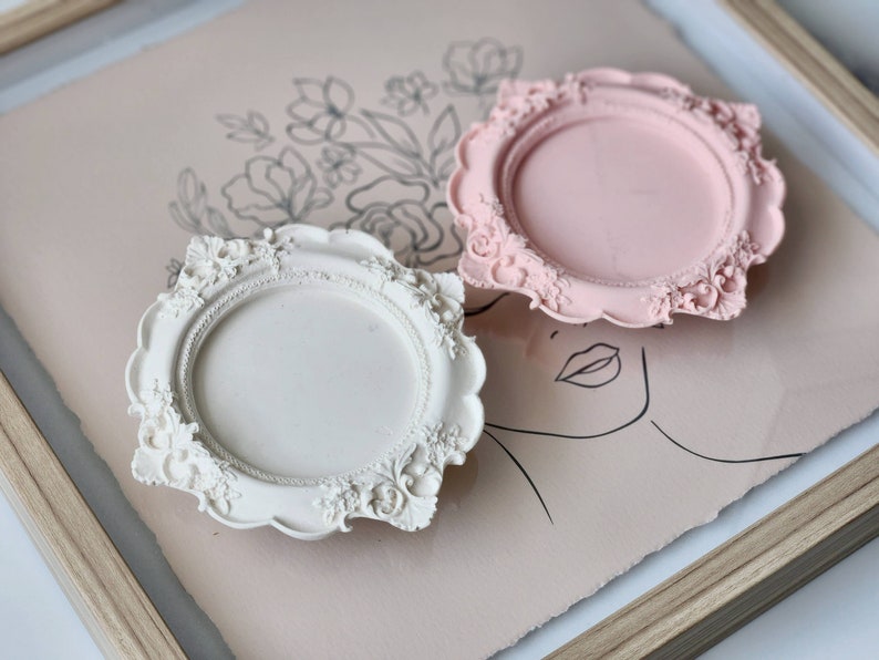 Puede incluir: Dos bandejas redondas decorativas, ornamentadas, blancas y rosas, con un marco y un dise&ntilde;o floral elevado y detallado.