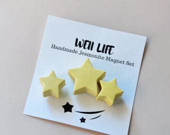 Puede incluir: Un conjunto de tres imanes en forma de estrella amarilla hechos de jesmonita. Los imanes están dispuestos en una tarjeta blanca con el texto "Well Life Handmade Jesmonite Magnet Set".