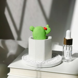 May include: A white ceramic planter with a green cactus and a red flower on top. The planter is on a white pedestal with a decorative design. A small glass bottle with a gold lid and a label that says "Lemon Crazy" is next to the planter.