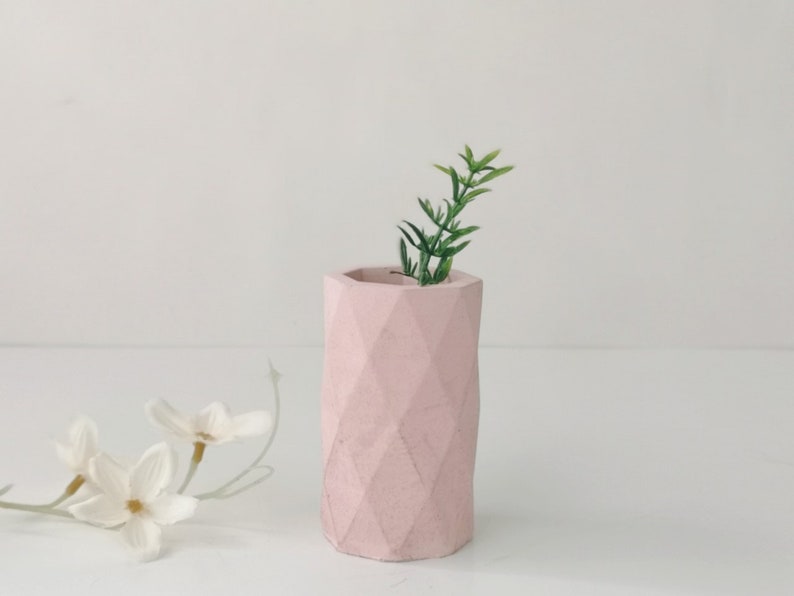 May include: A light pink, geometric-patterned vase with a small green plant inside. The vase has a cylindrical shape and a textured surface. The background is white, and there are white flowers in the foreground.