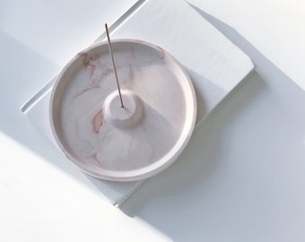 May include: A white ceramic incense holder with a marbled design and a small, raised center for holding incense sticks. The holder is on a white surface.