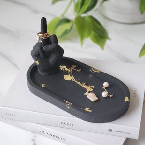 May include: Black ceramic ring dish with a gold flecked finish. The dish features a raised sculpture of a middle finger with rings on it. The dish is sitting on top of two books.