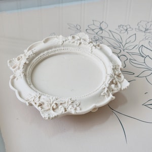 Puede incluir: Una bandeja decorativa de cer&aacute;mica blanca con un borde festoneado ornamentado y un centro circular. La bandeja tiene una est&eacute;tica vintage y antigua.