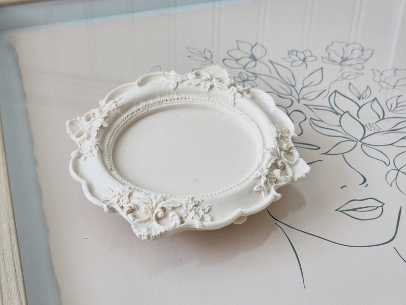 Puede incluir: Una bandeja decorativa de cer&aacute;mica blanca con un borde festoneado ornamentado y un centro circular. La bandeja tiene una est&eacute;tica vintage y antigua.