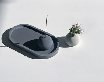 May include: A gray ceramic incense holder with a round top and a stick of incense. The holder is sitting on a gray oval tray. A small white ceramic vase with a sprig of white flowers is in the background.