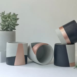 May include: Four concrete planters with geometric designs in black, white, and rose gold. The planters are arranged in a row on a white surface.