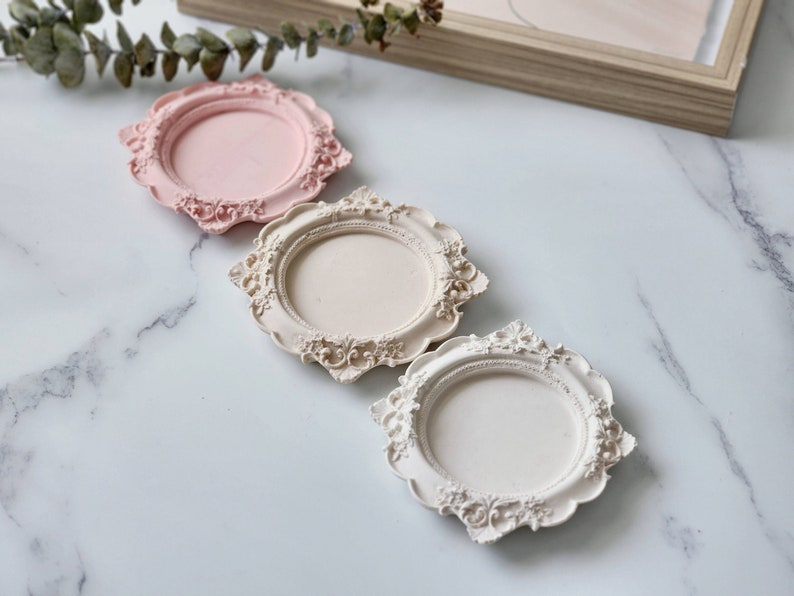 Puede incluir: Tres posavasos decorativos, ornamentados, redondos, blancos y rosados con un borde elevado. Los posavasos est&aacute;n hechos de un material liso y texturizado y tienen un aspecto vintage, antiguo.