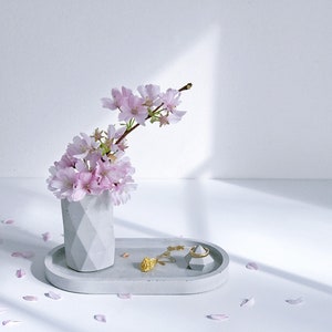 May include: A gray concrete tray with a geometric design holds a small vase with pink cherry blossoms and a ring holder. The tray is on a white surface with pink flower petals scattered around it.