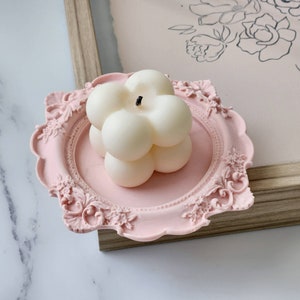 Puede incluir: Una vela blanca redonda de m&uacute;ltiples burbujas se asienta sobre una bandeja decorativa de color rosa adornada. La bandeja tiene un borde festoneado y dise&ntilde;os florales intrincados.