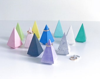 Puede incluir: Un conjunto de diez soportes de anillo geométricos en varios colores, incluyendo rosa, verde, azul, amarillo y blanco. Los soportes están hechos de hormigón y tienen forma de pirámide. Algunos de los soportes tienen anillos en ellos.