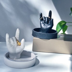 May include: Two ceramic hand ring holders, one black and one white, with a ring on each finger. The holders are on a beige surface.