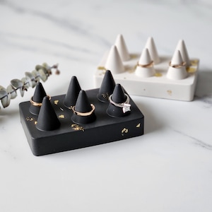 May include: A black and white ring holder with gold accents. The holder has multiple cone-shaped compartments for rings. The holder is made of concrete and has a minimalist design.