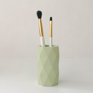 May include: A light green geometric makeup brush holder with two brushes. The brushes have gold and white handles and black bristles. The holder is cylindrical with a faceted design.