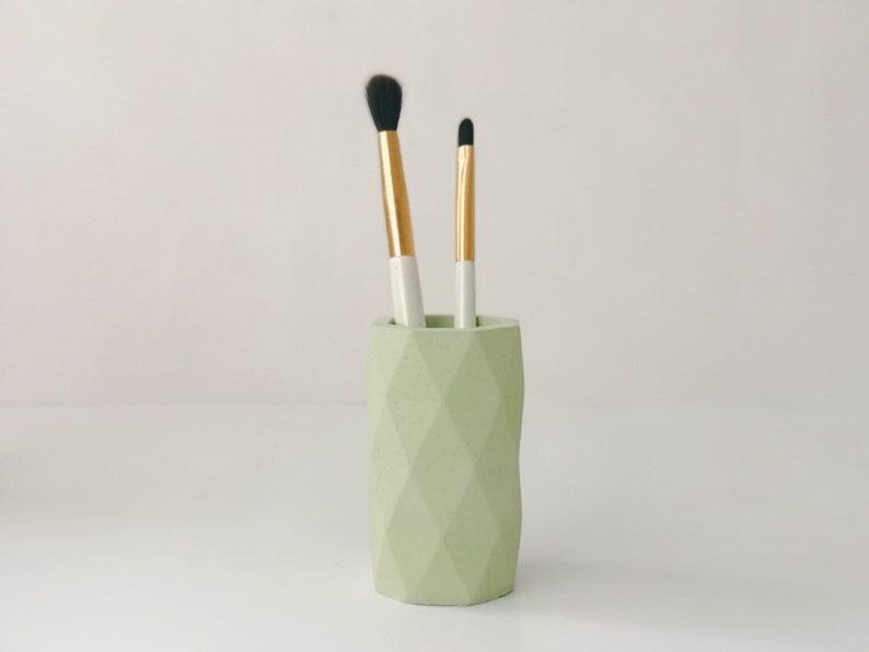 May include: A light green geometric makeup brush holder with two brushes. The brushes have gold and white handles and black bristles. The holder is cylindrical with a faceted design.