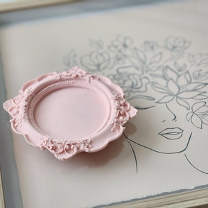 Puede incluir: Un marco decorativo rosa con un dise&ntilde;o ornamentado. El marco est&aacute; sobre una superficie rosa claro con un dibujo en blanco y negro de una cara de mujer con flores.
