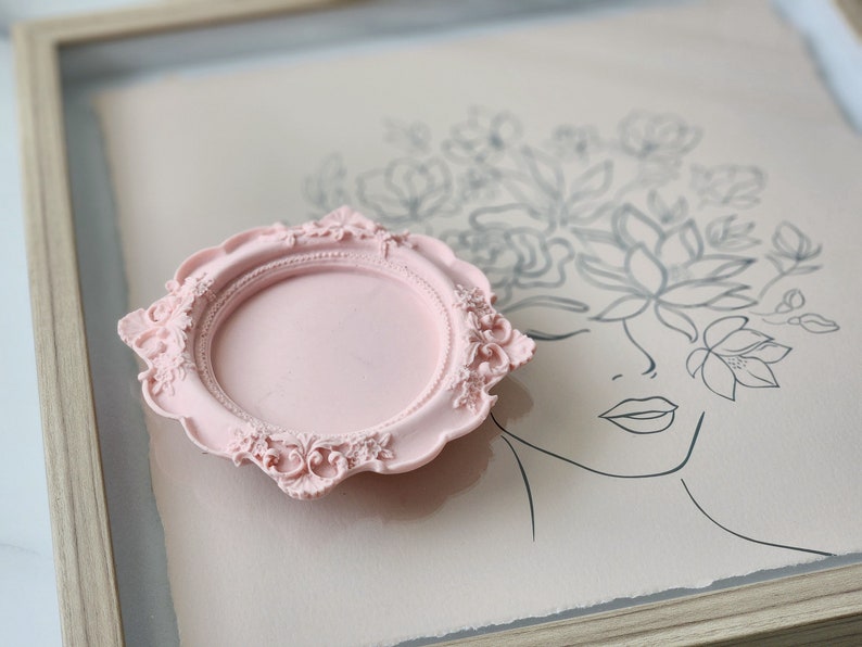 Puede incluir: Un marco decorativo rosa con un dise&ntilde;o ornamentado. El marco est&aacute; sobre una superficie rosa claro con un dibujo en blanco y negro de una cara de mujer con flores.
