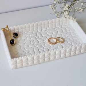 May include: White ceramic tray with a textured pebble design. The tray has a raised border with a beaded edge. Two gold rings and a pair of black earrings are on the tray.