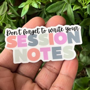 May include: A sticker with the text "Don't forget to write your SESSION NOTES." The text is in a mix of colors, including pink, gray, orange, and green. The sticker is held in a person's hand against a background of green leaves.