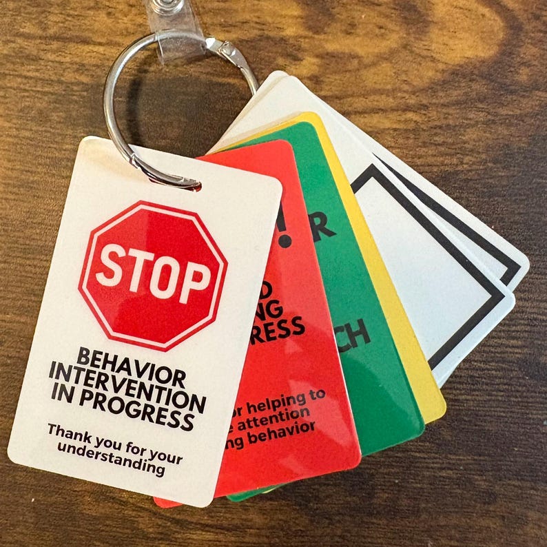Puede incluir: Un conjunto de llaveros coloridos con texto. El llavero principal es blanco con una se&ntilde;al de stop roja y las palabras "Behavior Intervention in Progress". Otros llaveros son rojos, verdes y amarillos, con texto adicional. Un anillo plateado sujeta los llaveros.