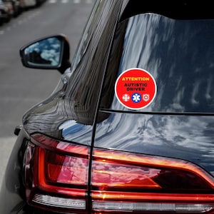 May include: A red and yellow round sticker on a black car reads "Attention Autistic Driver" with a red fire department symbol, a blue star of life, and a blue and white symbol.