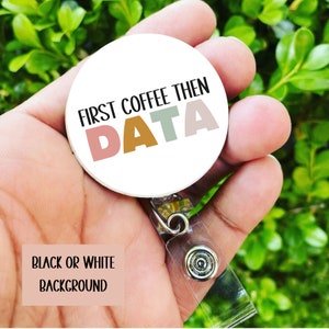 May include: A white round badge holder with a black and white background. The badge holder has a clear plastic retractable cord. The badge holder has the text "First Coffee Then Data" in black text with the word "Data" in colorful letters.