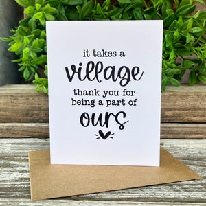 May include: White card with black text that reads "it takes a village thank you for being a part of ours" with a small heart symbol below the word "ours".