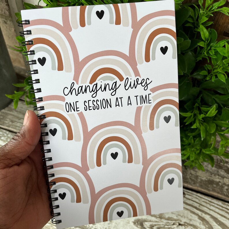 May include: A white spiral bound notebook with a rainbow pattern and the text "changing lives one session at a time".