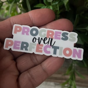 May include: A sticker with the words "PROGRESS over PERFECTION" in a mix of pastel colors. The word "PROGRESS" is in pink, blue, and gray block letters. The word "over" is in black cursive. The word "PERFECTION" is in pink, orange, and gray block letters.