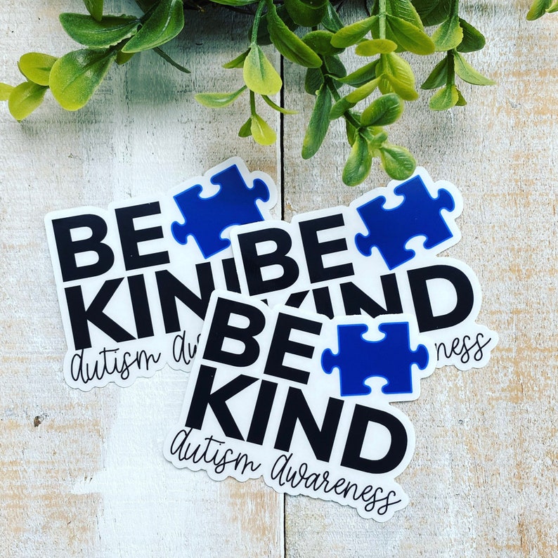 Puede incluir: Tres pegatinas blancas con texto negro que dice "Be Kind" y una pieza de rompecabezas azul. El texto debajo de las pegatinas dice "Conciencia del autismo".