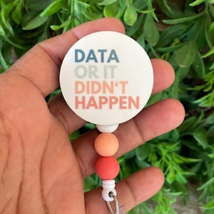 May include: A white round badge holder with the text "DATA OR IT DIDN'T HAPPEN" in blue, green, and pink lettering. The badge holder is attached to a retractable cord with three peach and coral colored beads.