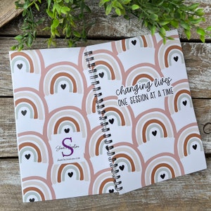 May include: A spiral bound notebook with a white background and a repeating pattern of brown, pink, and gray rainbows with black hearts. The text "changing lives one session at a time" is printed on the cover. The back cover has the text "Shane's Creations" and a website address.