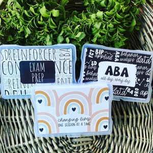 May include: Three clear plastic containers with colorful designs and text. The first container has black and white text that says "Exam Prep". The second container has black and white text that says "ABA all day, every day". The third container has a rainbow design and the text "changing lives one session at a time".