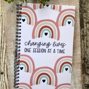 May include: A white spiral bound notebook with a rainbow pattern and the text "changing lives one session at a time".