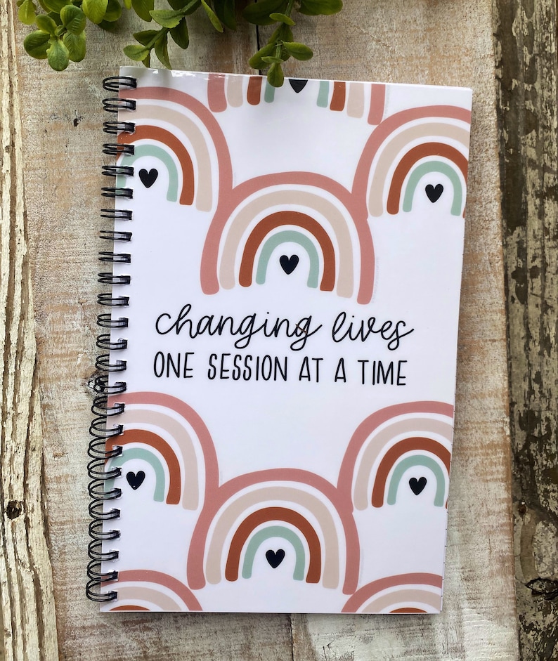 May include: A white spiral bound notebook with a rainbow pattern and the text "changing lives one session at a time".