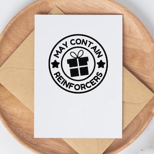 May include: A white greeting card with a black circular design that reads "MAY CONTAIN REINFORCERS" around a gift box icon. The card is on a brown envelope and a wooden tray.