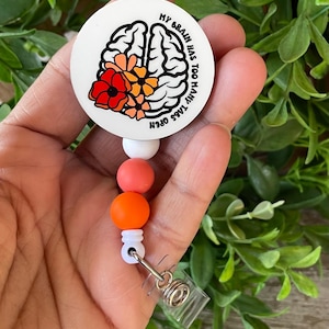 May include: A white round badge holder with a colorful brain graphic and the text "My brain has too many tabs open". The badge holder has a retractable cord with orange, pink, and white beads.