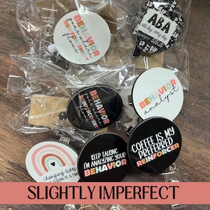 May include: A collection of badge reels with various designs and phrases. The reels feature phrases like "Behavior analyst," "Coffee is my preferred reinforcer," and "Behavior goes where reinforcement flows." The background is a wooden surface.