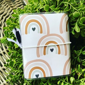 May include: A white notebook cover with a repeating pattern of brown, beige, and white rainbows with black heart accents.