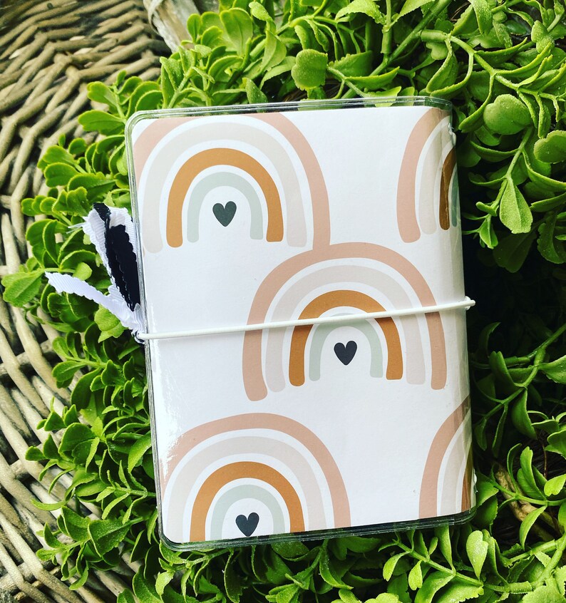 May include: A white notebook cover with a repeating pattern of brown, beige, and white rainbows with black heart accents.