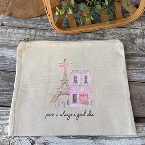 May include: A beige canvas zippered pouch featuring a watercolor illustration of the Eiffel Tower and a pink cafe. The phrase "paris is always a good idea" is written in black script below the illustration. The pouch has a silver zipper.
