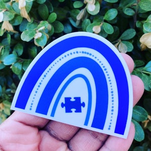 May include: A blue and white rainbow sticker with a blue puzzle piece in the center. The rainbow has a white outline and is decorated with white dots.